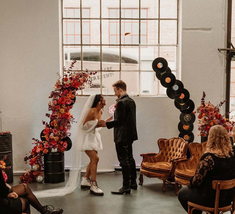 Bride in off shoulder tulle short wedding dress with church length veil and white platform Dr Martens wedding shoes laughing with groom in wet look black grooms suit with leopard print shirt and pink and orange boutonniere standing by vinyl wedding arch 