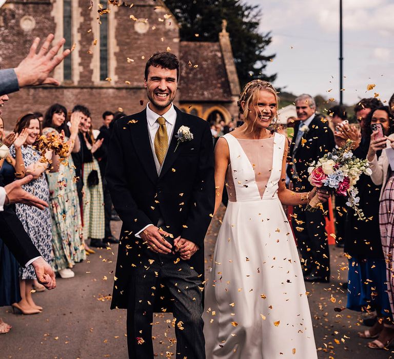 Groom in morning suit with yellow tie walking hand in hand with the bride to colourful confetti exit 