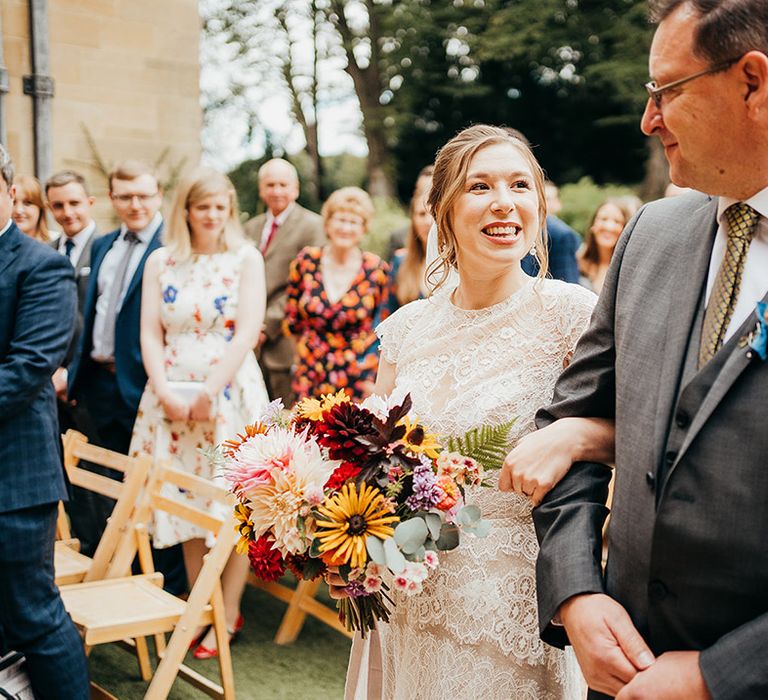 Father of the bride in grey suit linked arms with the bride as they walk down the aisle together 