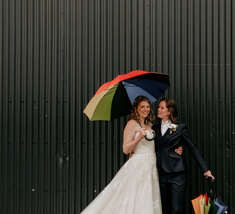 Lesbian bride in a strapless lace wedding dress and navy suit holding rainbow pride umbrellas 