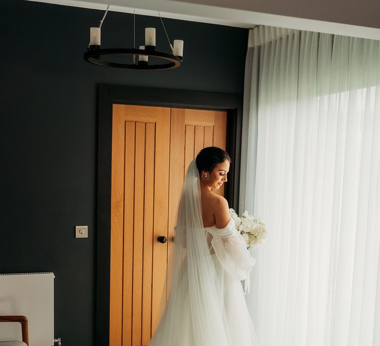 Bride in a white tulle off-the-shoulder wedding dress with bridal veil and bridal updo holds white rose wedding bouquet