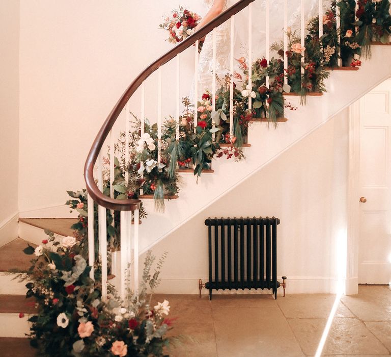 The bride and bridesmaids walk down the stairs together covered in festive seasonal winter flowers 