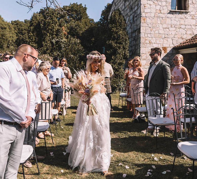 Bride holding pampas grass wedding decor in an off the shoulder boho wedding dress walking down the aisle by herself 