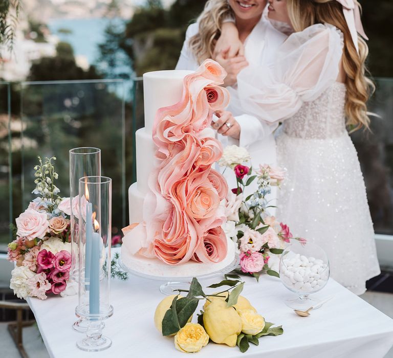 Two brides embrace as they cut their wedding cake together which is decorated with pink ribbon 