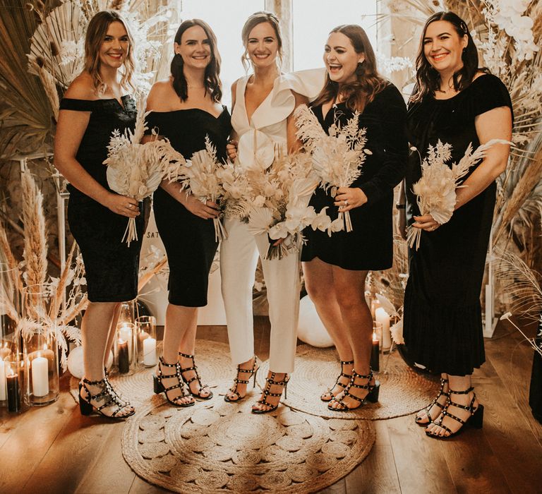 Bride in white jumpsuit and bridesmaids in black dresses for stylish micro wedding with dried flowers 