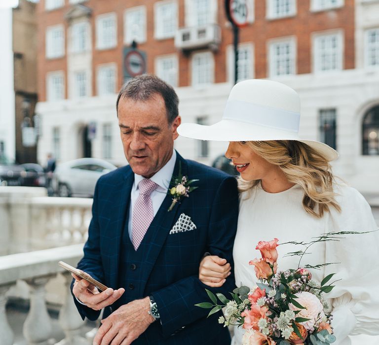 Bride hooked arms with the father of the bride wearing a white wedding hat, peach roses and coral lipstick