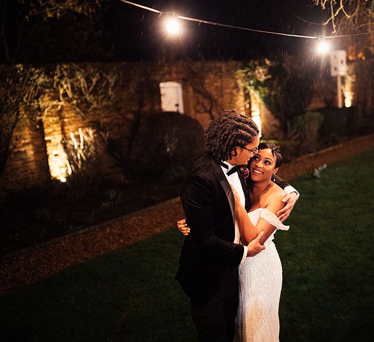 A bride and groom embrace outside at night time. The bride has short cropped hair.