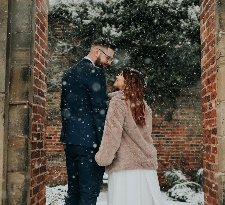 Bride in fur coat holds hands with groom in the snow in Cannon Hall gardens 