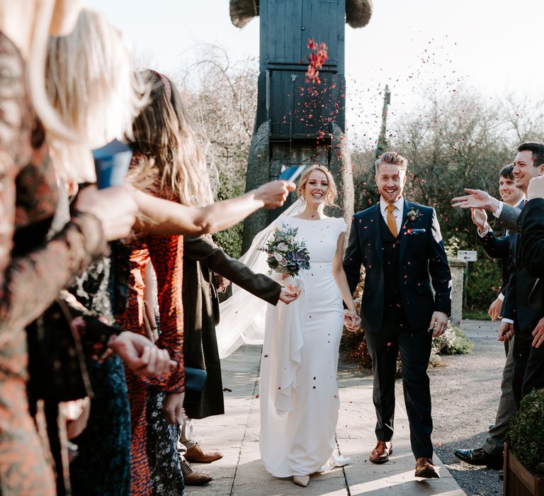 Confetti moment with bride in a fitted pronovnas wedding dress holding hands with her groom in a navy suit 