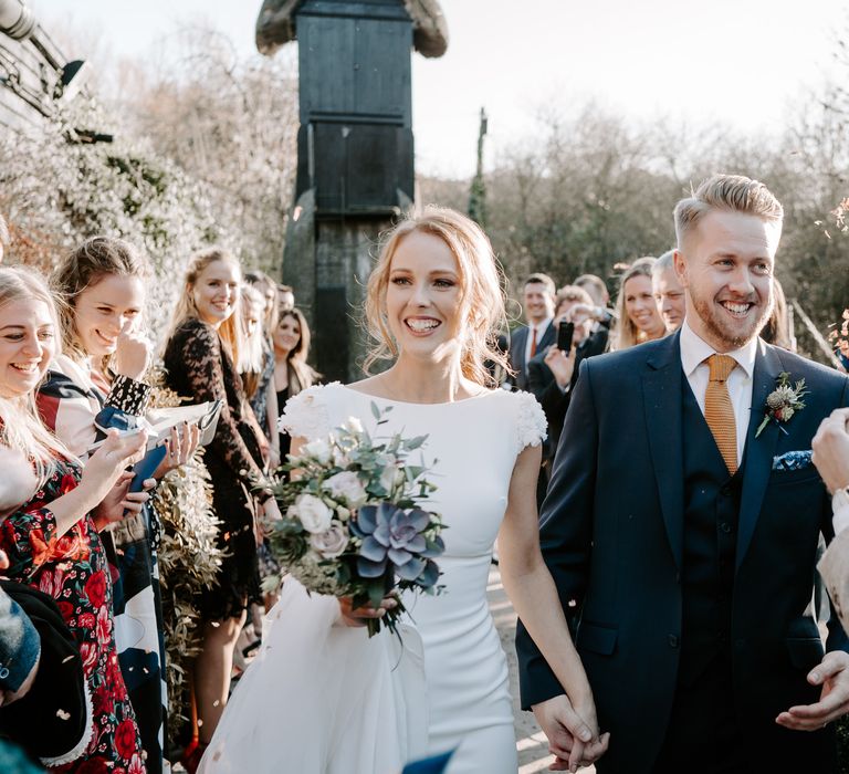 Confetti moment with red headed bride in a fitted wedding dress and cathedral length veil holding a succulent bouquet and her grooms hand in a navy three-piece suit