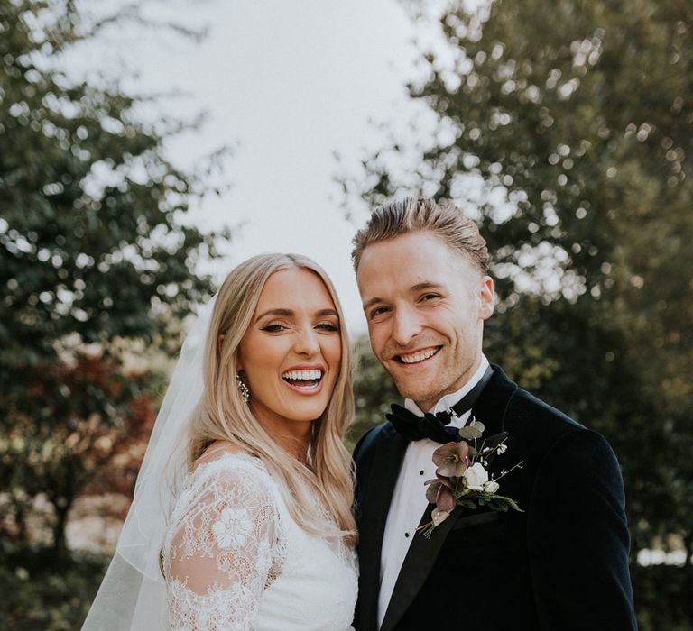 Bride in white Charlie Brear dress, white lace Augusta Jones top and full length veil standing with groom in black velvet Hugo Boss suit in the grounds at Caswell House wedding