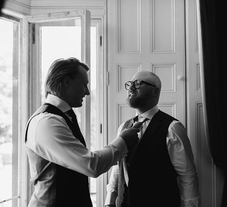 Groomsman in white shirt and waistcoat ties grooms tie by window 