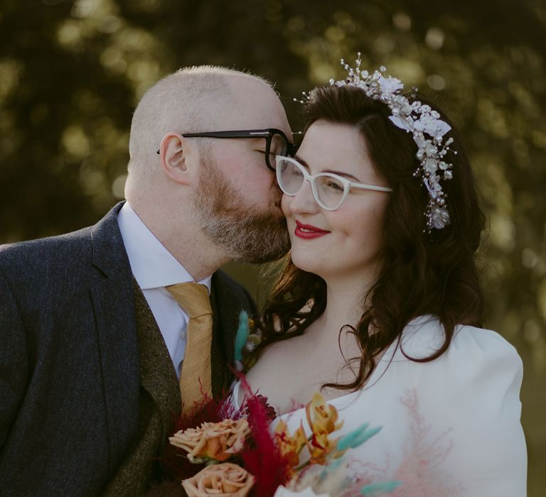 Groom in navy suit and yellow tie kisses bride in white cat eye glasses and bridal headband on the cheek as she holds white and pink rose and pampas grass bouquet