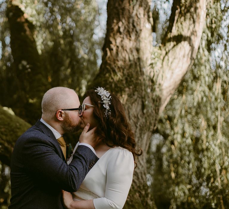 Groom in navy suit and yellow tie kisses bride in white cat eye glasses and bridal headband holding white and pink rose and pampas grass bouquet