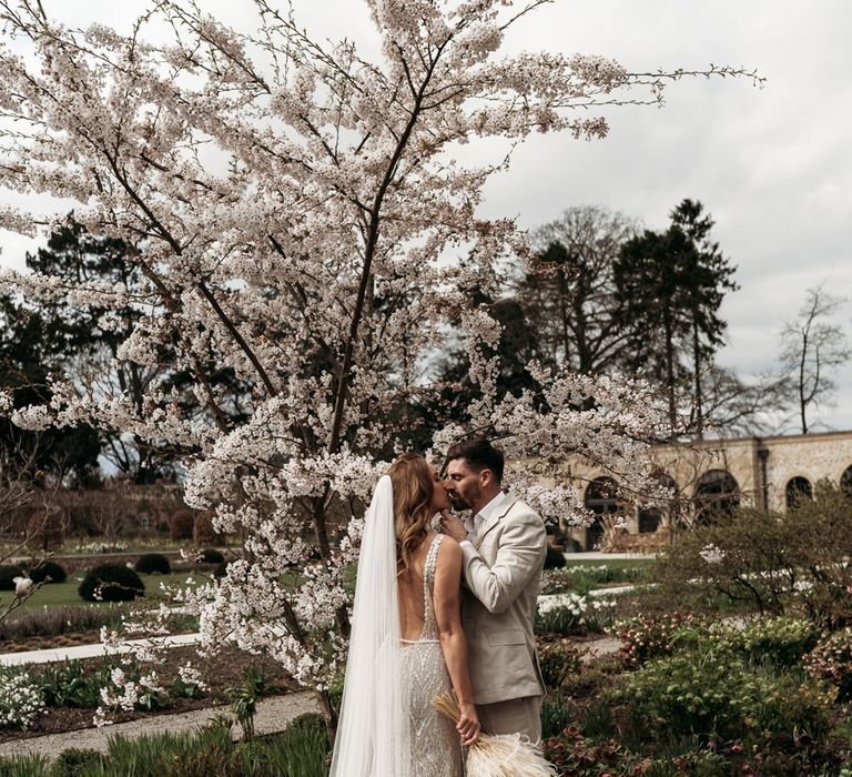 Bride in lace wedding dress holding a dried flower bouquet kissing her husband in beige suit under a blossom tree 