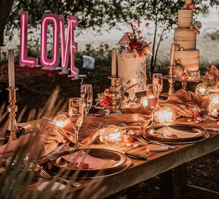 Outdoor wedding tablescape at festival themed wedding with candles, neon sign, three tiered naked wedding cake and tropical print champagne flutes 