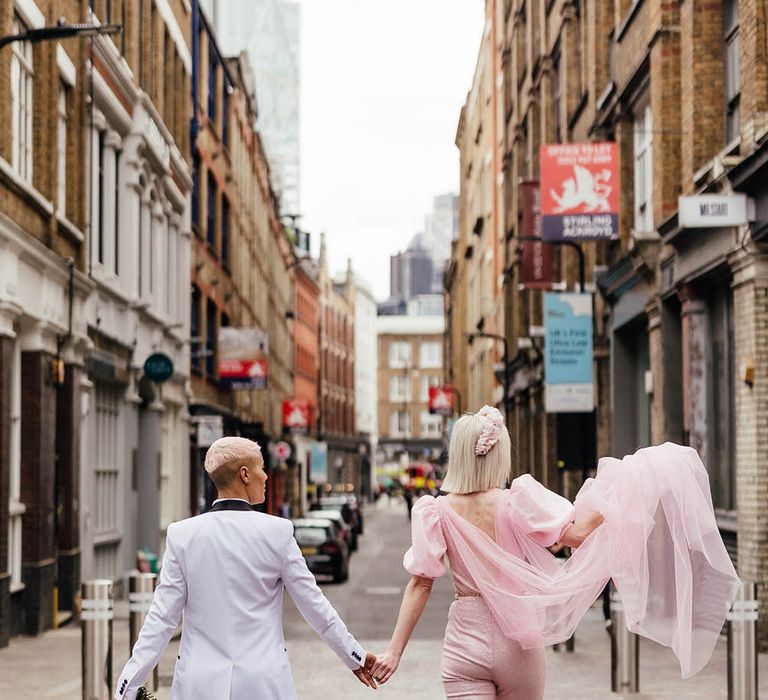 Two brides walking through Shoreditch holding hands in a pink jumpsuit with Watteau train and white tuxedo jacket 