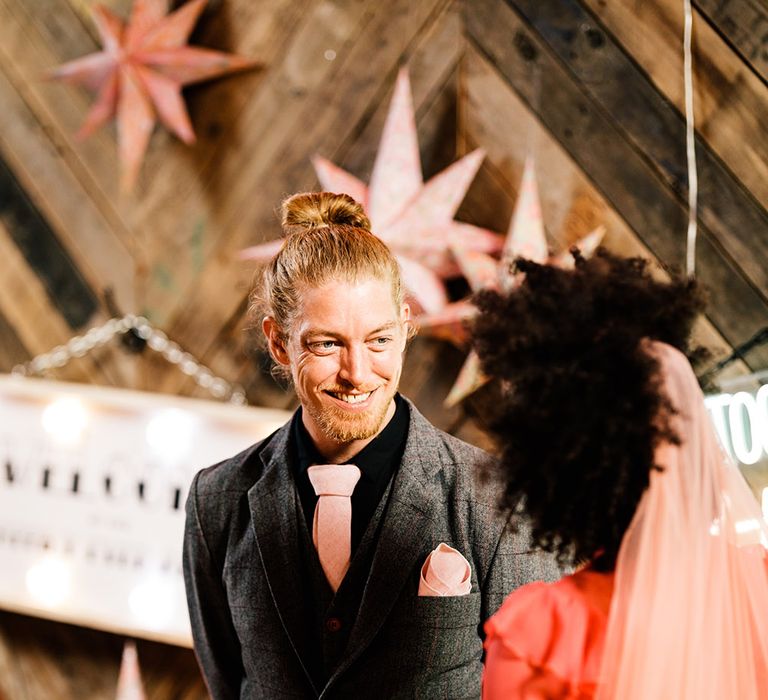 Groom with a top knot in a black shirt, pink tie and grey check suit looking at his bride during the intimate wedding ceremony 