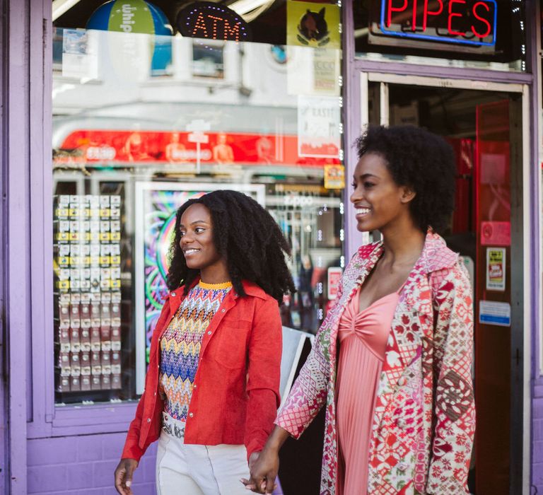 Bride in white jeans, patterned top and orange jacket holding hands with her bride-to-be in a coral dress and patterned longline coat 