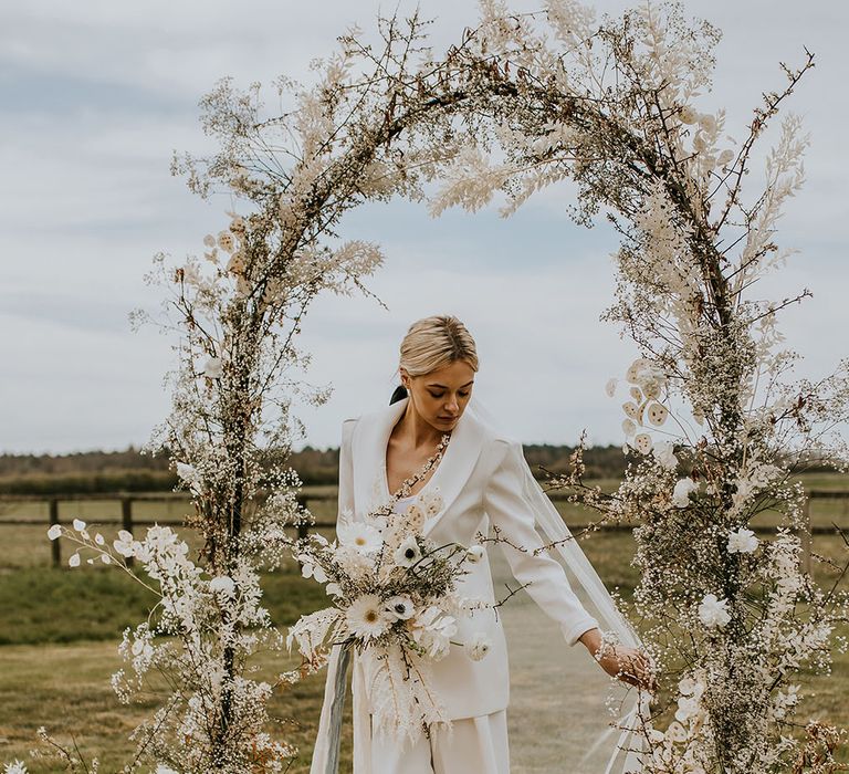 Bride in a white suit standing under an all-white flower arch at bohemian luxe wedding 