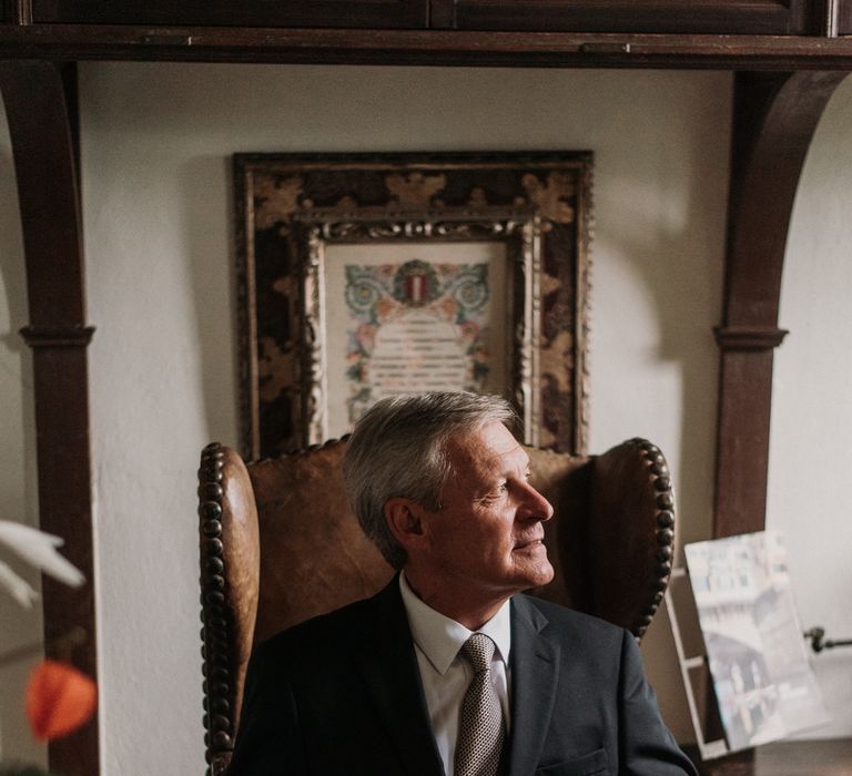 Groom sits in chair as he looks to the side whilst wearing black jacket, white shirt and silver patterned tie