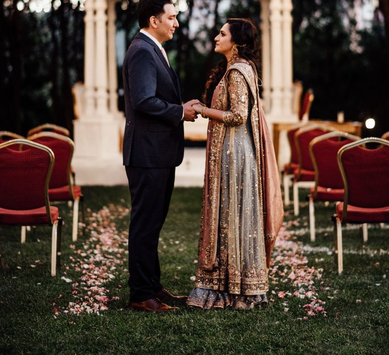 Bride & groom stand between chairs of petal line aisle as floral covered bandstand can be seen in the background