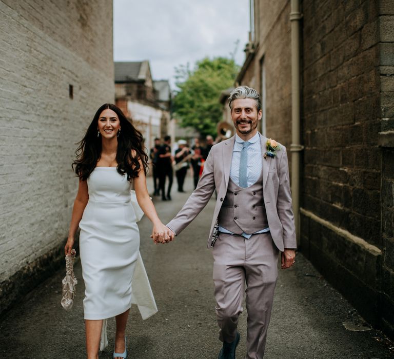 Bride in white strapless Rebecca Vallance Dress holding ivory bridal bag walks through the streets of Harrogate holding hands with groom in brown Moss Bros suit and blue tie whilst marching band follows them