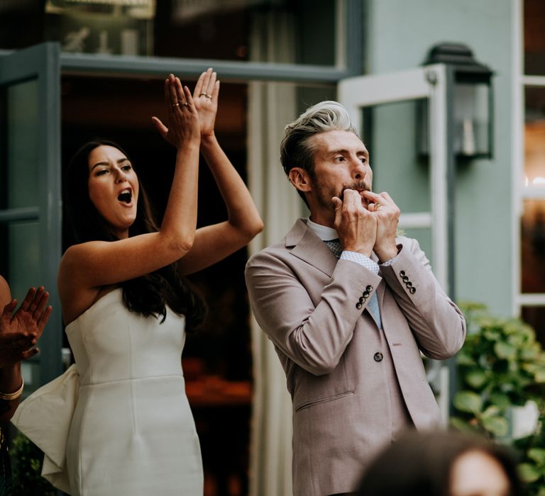 Bride in white strapless Rebecca Vallance Dress claps whilst groom in light brown Moss bors suit and blue tie whistles with both fingers during courtyard wedding reception at Hotel du Vin Harrogate