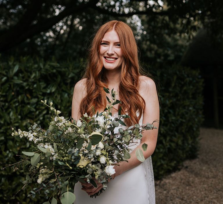 Beautiful bride with long red hair wearing a sating slip Grace Loves Lace wedding dress and holding a giant green and white wedding bouquet 