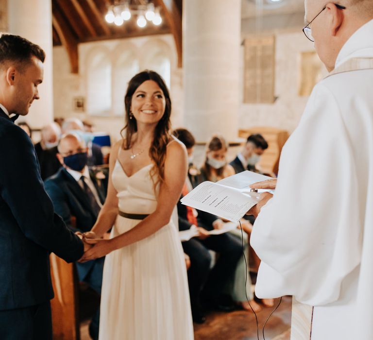 Bride in white cami wedding dress with gold ribbon waistband smiles at the vicar whilst holding hands with groom in blue suit during wedding ceremony