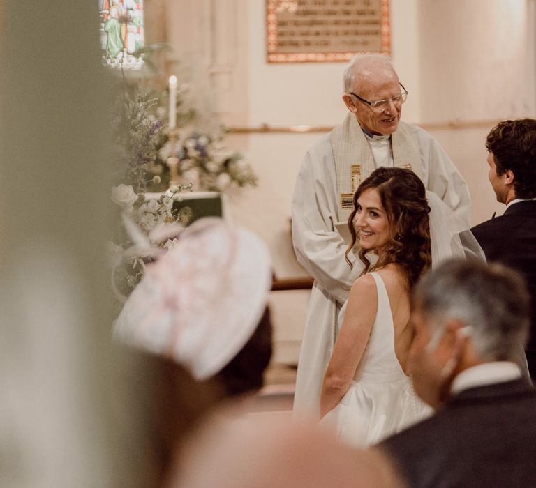 Bride in low back white Elbeth Gillis gown, long curled hair and veil smiles back at the congregation whilst kneeling at the alter during church wedding ceremony