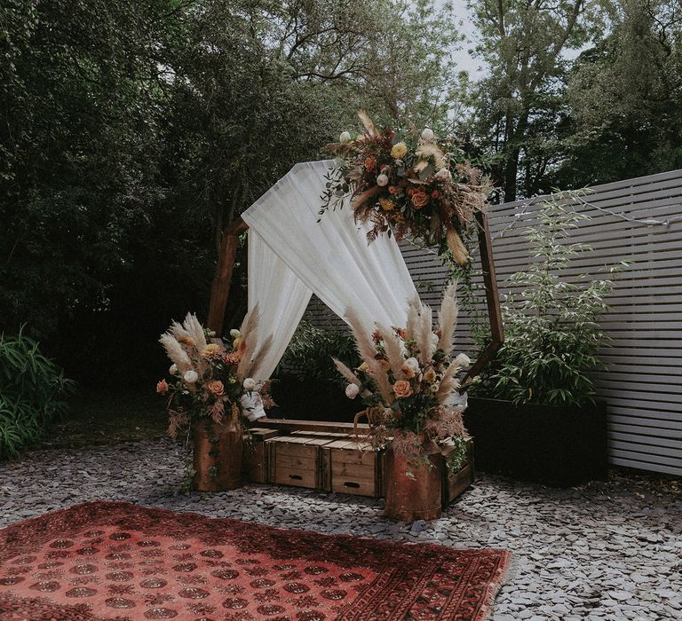 Wooden hexagonal wedding arch with white tulle drapery, floral and pampas grass installations and crates on bed of flint with Persian rug at garden wedding with burnt orange wedding theme 