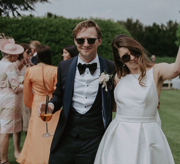 Bride & groom wear sunglasses outdoors on their wedding day as they celebrate with friends