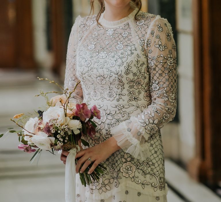 Bride smiles brightly as she wears Needle And Thread wedding dress complete with floral bouquet in her hands and loose up-do 