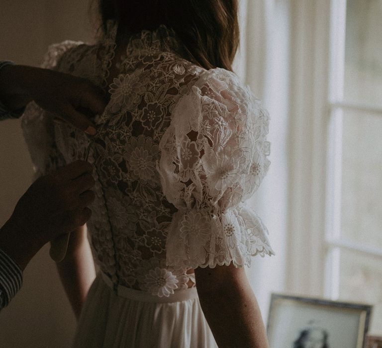 Bride in lace puffed sleeved Daalarna wedding dress has the back buttoned up as she stands by window before Surrey summer wedding