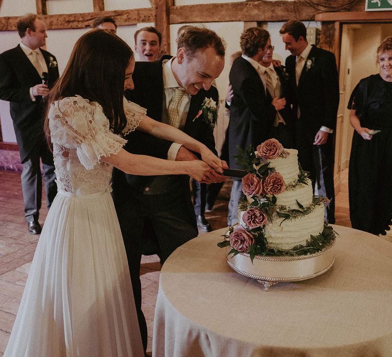 Bride in white lace puffed sleeve Daalarna wedding dress and groom in morning coat and olive leaf print tie cut their white three tier buttercream wedding cake during summer wedding at Loseley Park in Surrey 