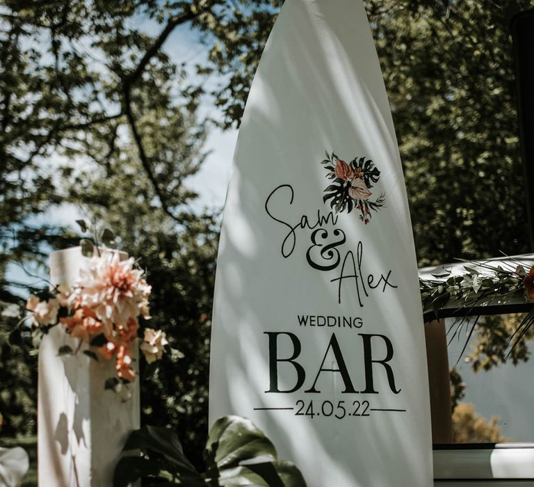 Large white surfboard sign reading 'Sam & Alex wedding bar 24.05.22' resting next to two tier white rectangular column cake and palm leaf for birds of paradise wedding inspiration