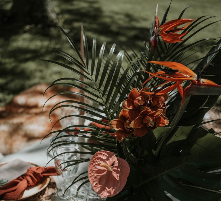 Tropical orange flowers and palm leaves for boho Hawaii wedding breakfast with red napkins, gold tableware and clear glasses for birds of paradise wedding inspiration