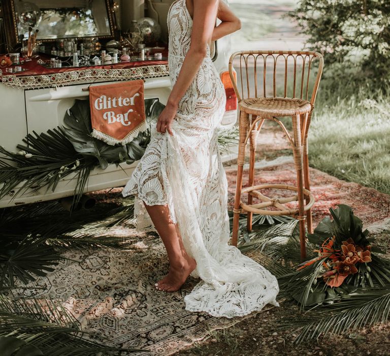 Bride with blonde beach waves holds up skirt of white lace boho wedding dress as she stands in front of glitter bar in the back of white campervan decorated with tropical flowers for birds of paradise wedding inspiration