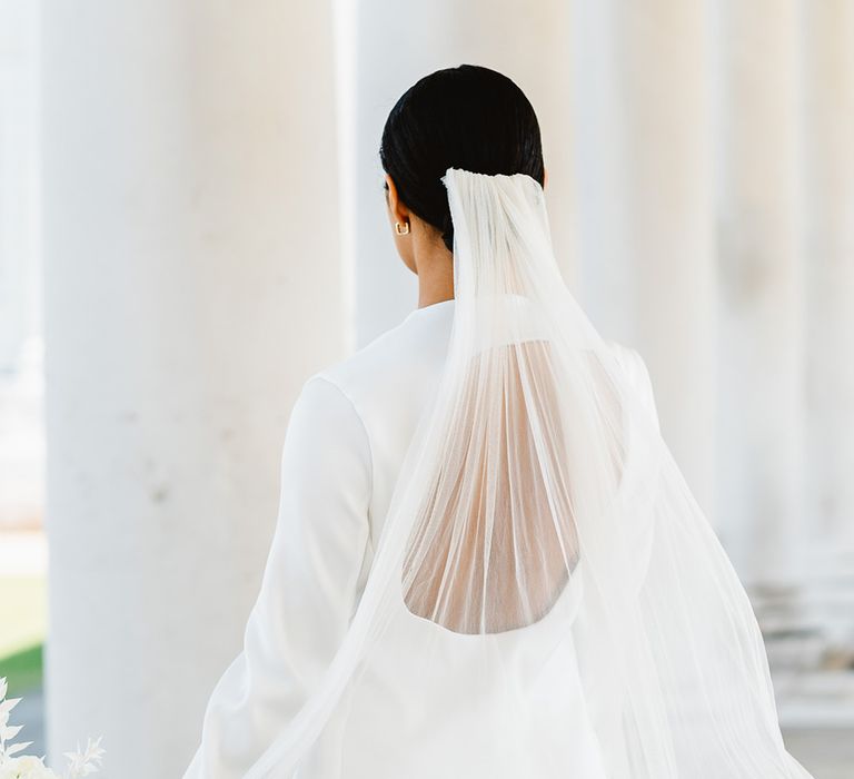 A bride stands with her back to the camera wearing a bespoke backless suit. Her veil is long and she holds black and white wedding flowers.