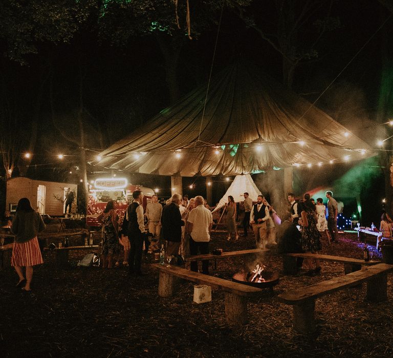 Wedding guests outdoors surrounded by fairy lights, fire pit and coffee van