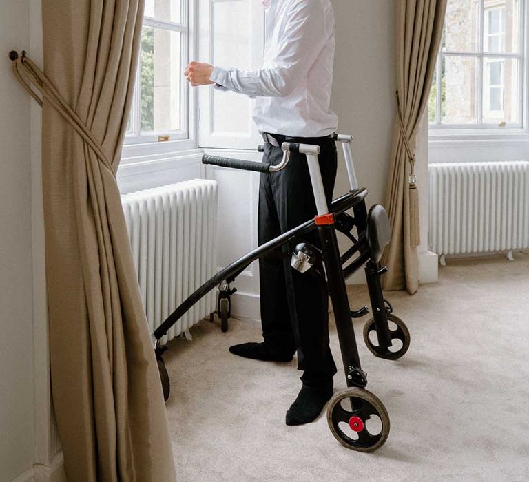 Disabled groom in a tuxedo standing at the window of the country house wedding venue with his walking frame 