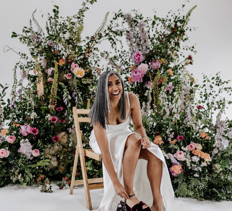 Bride in satin Halfpenny London wedding dress sat on wooden folding chair puts on burgundy velvet block heels in front of multicoloured floral installation for wedding at Loft Studios London