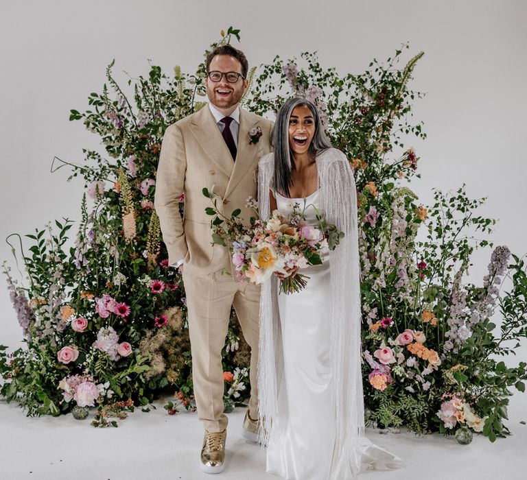 Bride in satin Halfpenny London wedding dress and tasselled bridal cape holding multicoloured bridal bouquet stands with groom in linen suit and gold Nike Air Force 1 trainers in front of floral installation for wedding at Loft Studios London