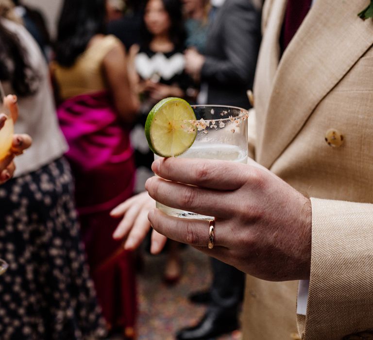 Groom in linen suit holds drink with salted rim and lime garnish during wedding reception at Loft Studios London