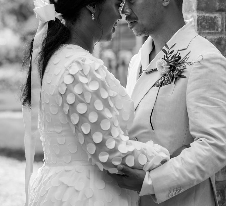 Groom in double breasted linen suit with floral buttonhole stands holding arms of bride in long mesh sleeve spotted Vagabond wedding dress and bridal hair bow outside at Wasing Park wedding