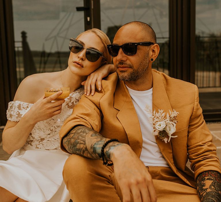 Stylish bride and groom in a Jesus Peiro wedding dress with lace off the shoulder bodice and mustard suit wearing sunglasses 