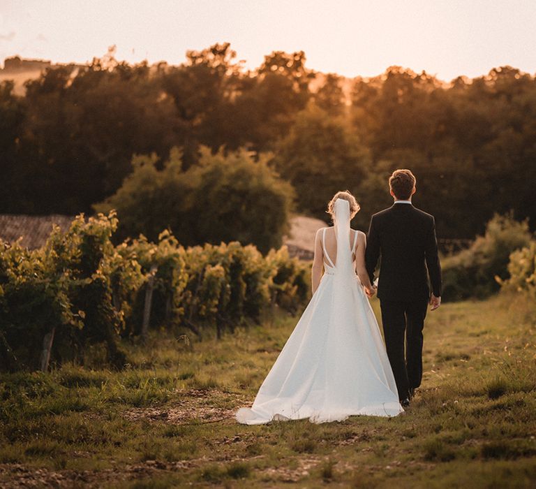 Wedding photography of the bride and groom holding hands during golden hour walking through a vineyard 