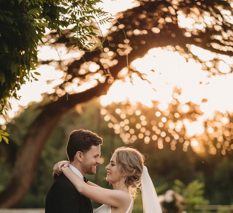 Bride in a princess wedding dress and cathedral length veil embracing her groom in a tuxedo during golden hour 