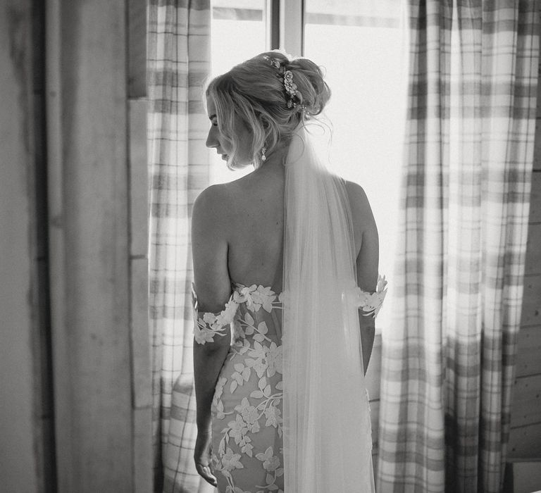 Bride in off the should Enzoani wedding dress, curled updo and veil stands by window before tipi wedding at Inkersall Grange Farm